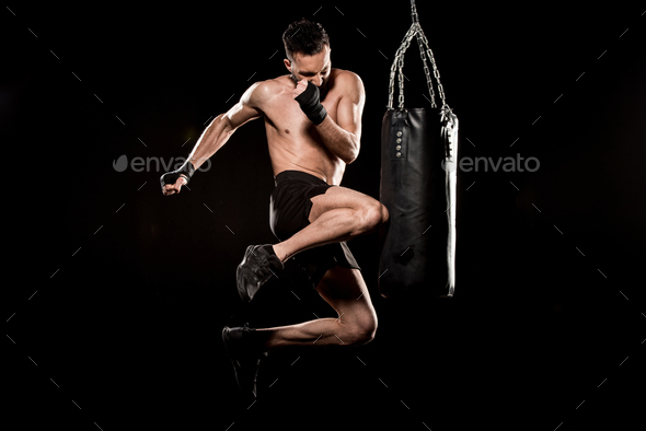 shortless athlete performing flying kick near punching bag isolated on black
