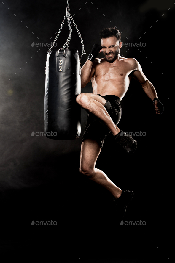 athletic boxer performing flying kick near punching bag on black with smoke