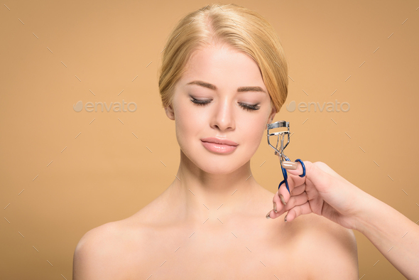 stylist holding tongs and curling eyelashes to beautiful blonde woman isolated on beige