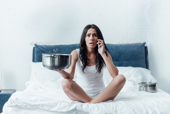 Shocked woman talking on smartphone and holding pot under leaking ceiling in bedroom
