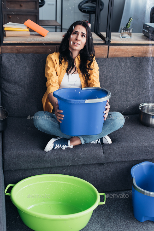 Upset woman with buckets and basin looking at leaking ceiling