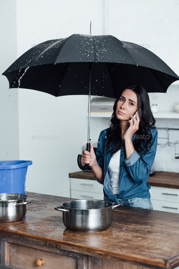 Sad woman talking on smartphone and holding umbrella under leaking ceiling in kitchen