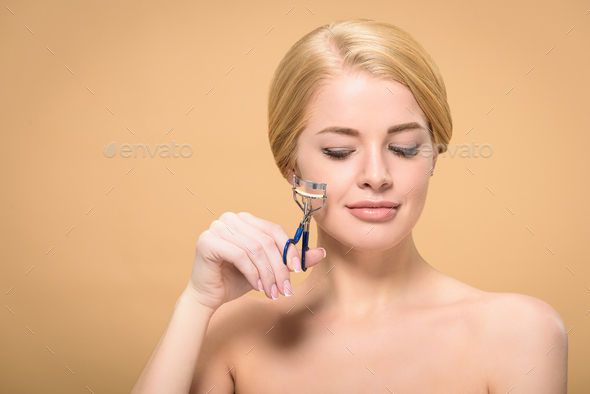 beautiful smiling naked girl holding curling tongs and looking down isolated on beige