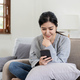 Image of smiling nice asian woman using cellphone while sitting on sofa - PhotoDune Item for Sale