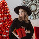 woman in a black dress and hat with a New Year&#39;s gift on the background of a red Christmas tree - PhotoDune Item for Sale