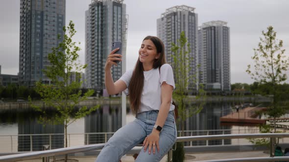 A Young Brunette Girl Sits in a Park on a Railing and Communicates By Video Call on the Phone