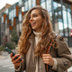 Smiling curly woman wearing warm coat walking down the street and using her phone - PhotoDune Item for Sale