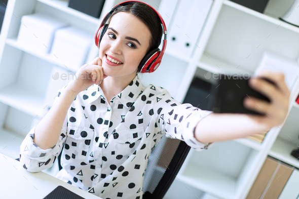 Beautiful young girl sitting in headphones at desk in office and taking pictures of herself on the