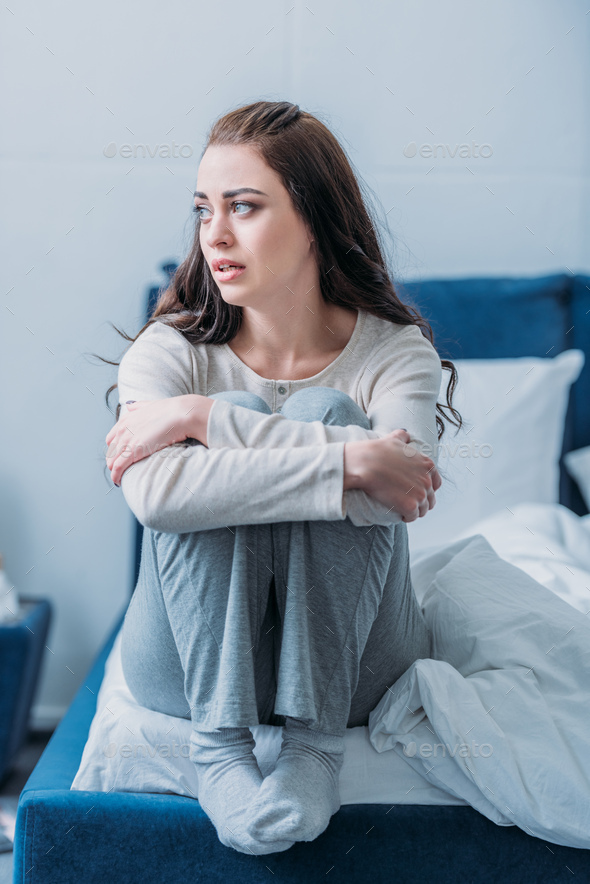 upset woman hugging knees, sitting on bed at home and looking away