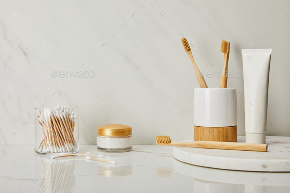 toothpaste in tube, holder, bamboo toothbrushes, cosmetic cream and ear sticks in glass on white