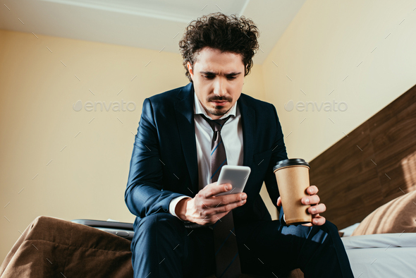 upset businessman using smartphone and holding disposable cup of coffee in hotel room