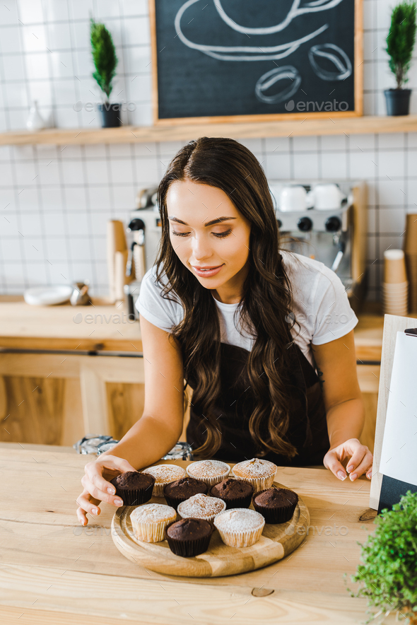 attractive brunette waitress standing behind bar counter and putting cupcakes on wooden tray in