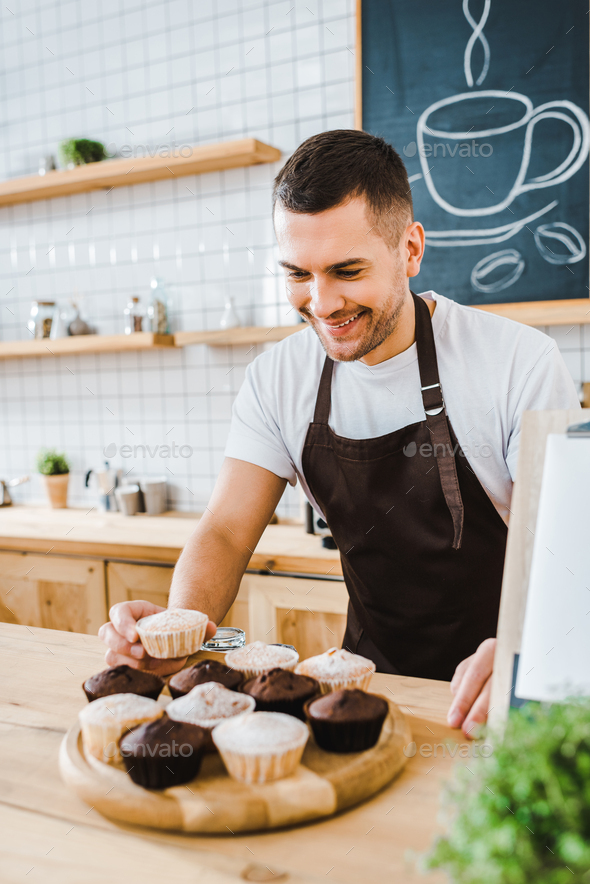 handsome waiter standing behind bar counter and putting cupcakes on wooden tray in coffee house