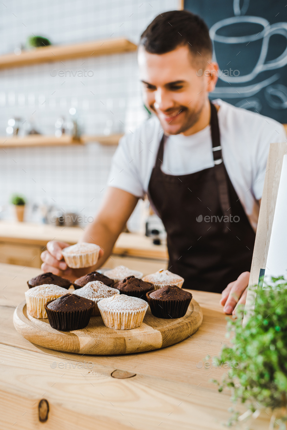 selective focus of barista standing behind bar counter and putting cupcakes on wooden tray in coffee
