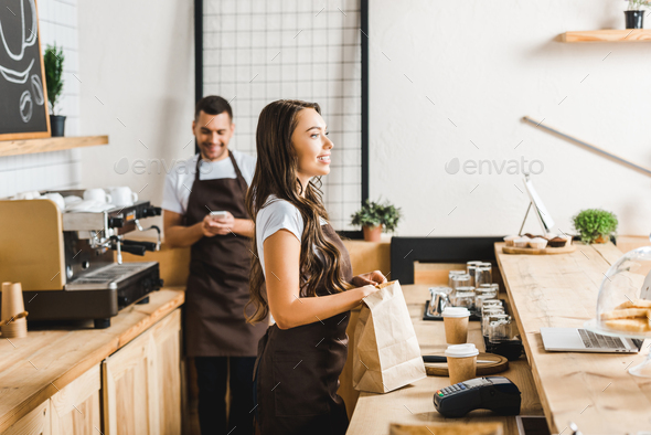 selective focus of attractive cashier with paper bag and barista with smartphone standing behind bar