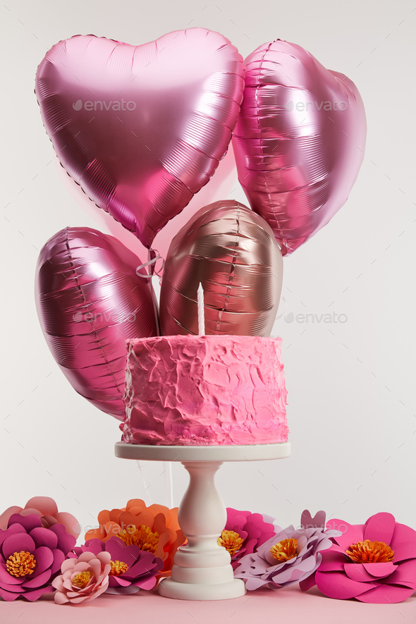 tasty pink birthday cake with candle on cake stand near paper flowers