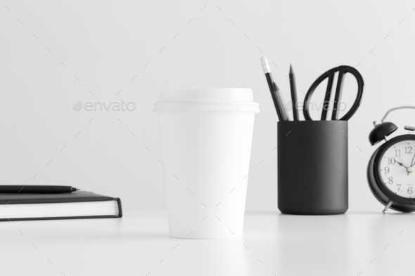 Coffee paper cup mockup with a notebook, clock and workspace accessories on a white table.