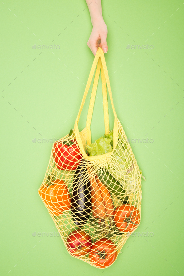 partial view of woman holding string bag with vegetables on light green