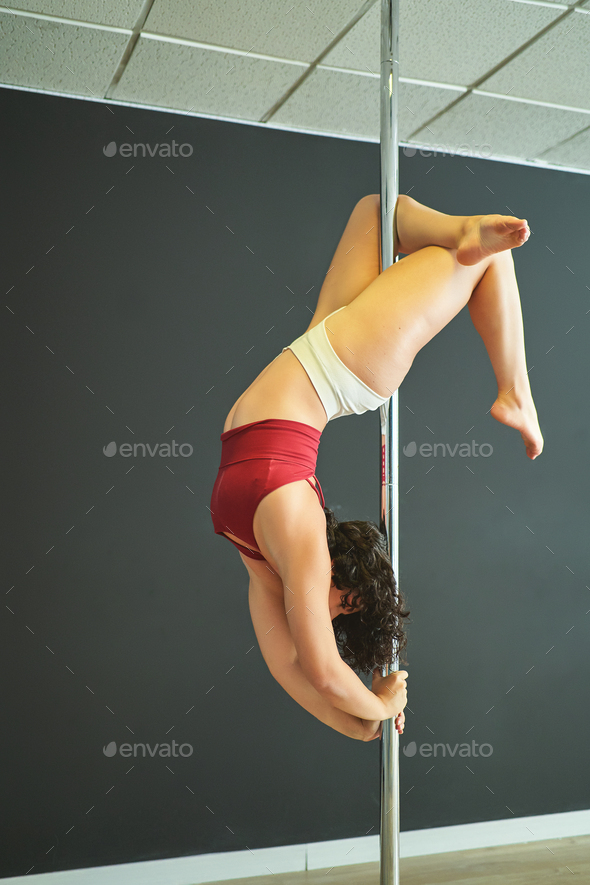 Young woman doing upside down pole dance exercises in a rehearsal room.