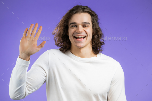 Young friendly man waving hand - hello. Greeting, say Hi to camera. Guy on violet studio background.