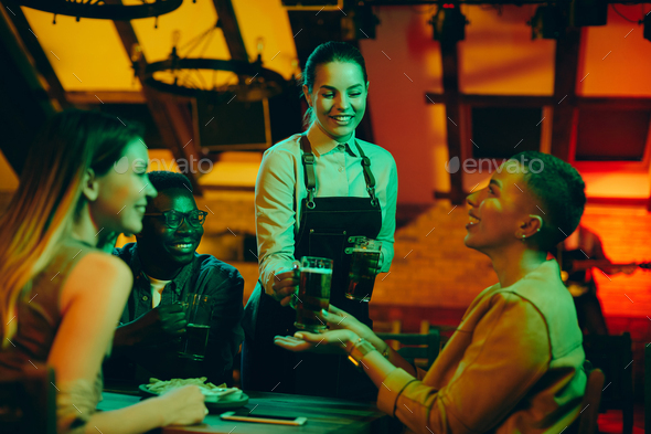 Young happy waitress serving beer to group of friends in a pub.
