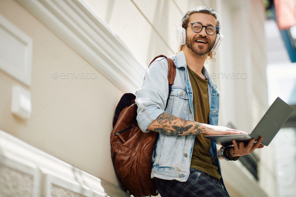 Happy male student with headphones using laptop in university hallway.