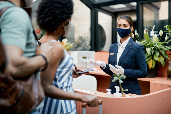 Happy receptionist with face mask talking to hotel guests during their check-in.
