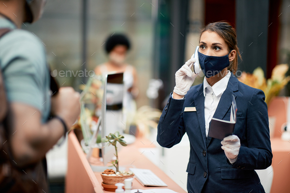Female receptionist with protective face mask talking on the phone during guest check-in i hotel.