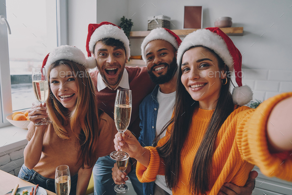 Beautiful people in Christmas hats holding champagne flutes while making selfie at home - Stock Photo - Images