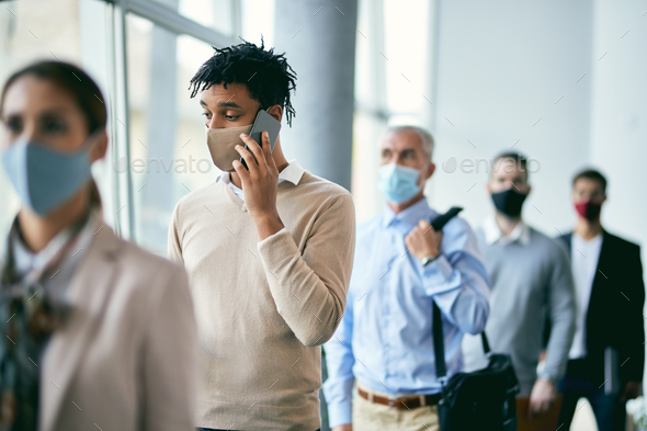 Young black businessman with face mask talking on the phone while waiting in line for job interview.