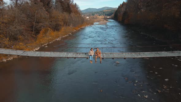Drone Selfie Footage By Couple Happy Hikers Sitting on Wooden Suspension Bridge Over Mountain River alt