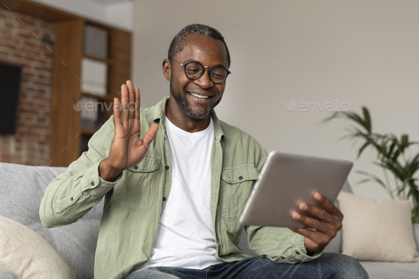 Glad happy middle aged black guy in glasses waving hand at digital tablet webcam, say hello, hi