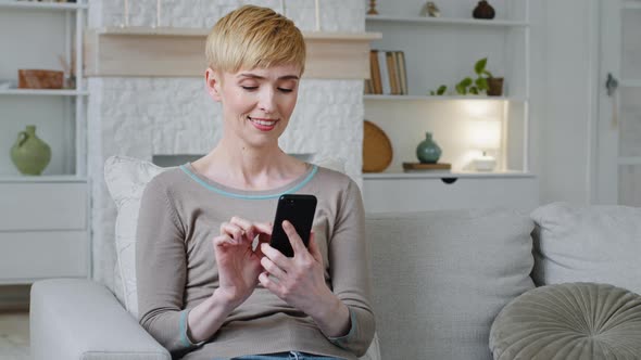 Happy Relaxed Woman Sitting on Sofa Holding Smartphone Looking at Cellphone Screen Surfing Social alt