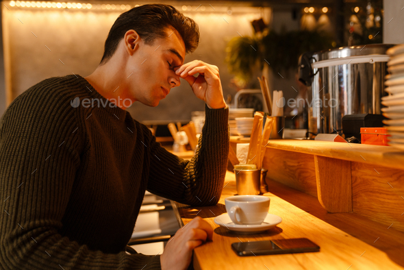 Young white man rubbing his eyes while drinking coffee in cafe