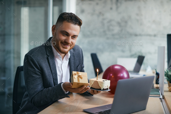 Happy businessman holding Birthday present and cake while making video call over laptop in office.