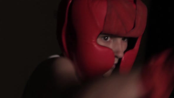 Young guy boxer practices punches in a dark room. Boxing protective helmet and hand bandages