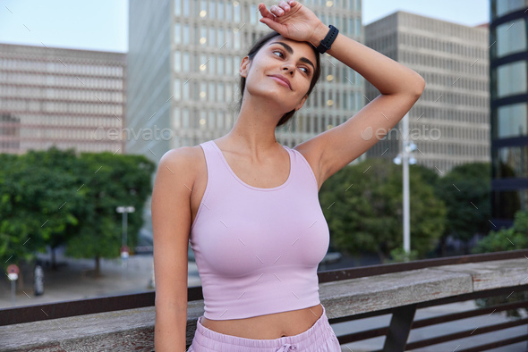 Tired sportswoman wipes forehead after hard fitness training wears cropped top smartwatch looks thou