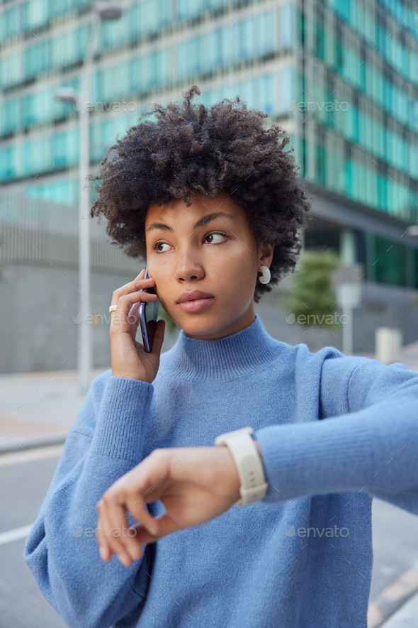 Photo of good looking curly haired Afro American woman calls friend waits for meeting checks time on