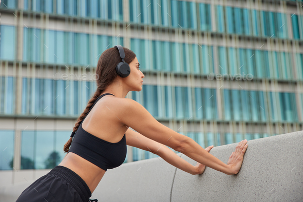 Sporty slim woman does push up exercises leans on concrrete wall wears cropped top stereo headphones