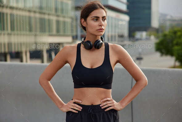 Thoughtful fit sportswoman keeps hands on waist looks away wears black cropped top and shorts headph