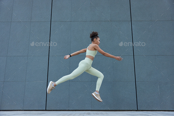 Photo of active slim woman in tracksuit jumps high against grey wall with blank space for your adver