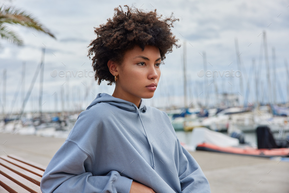 Photo of beautful thoughtful young Afro American woman dressed in hoodie poses on bench at pier bing - Stock Photo - Images