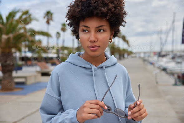 Outdoor shot of beautiful Afro American woman with curly hair holds sunglasses dressed in hoodie wal