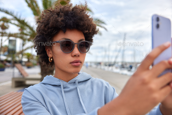 Photo of serious Afro American woman wears sunglasses hoodie poses at smartphone camera makes selfie