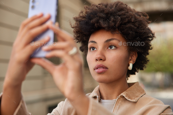 Serious female tourist uses smartphone to take selfie poses at city street  wears beige jacket focuse Stock Photo by wayhomestudioo