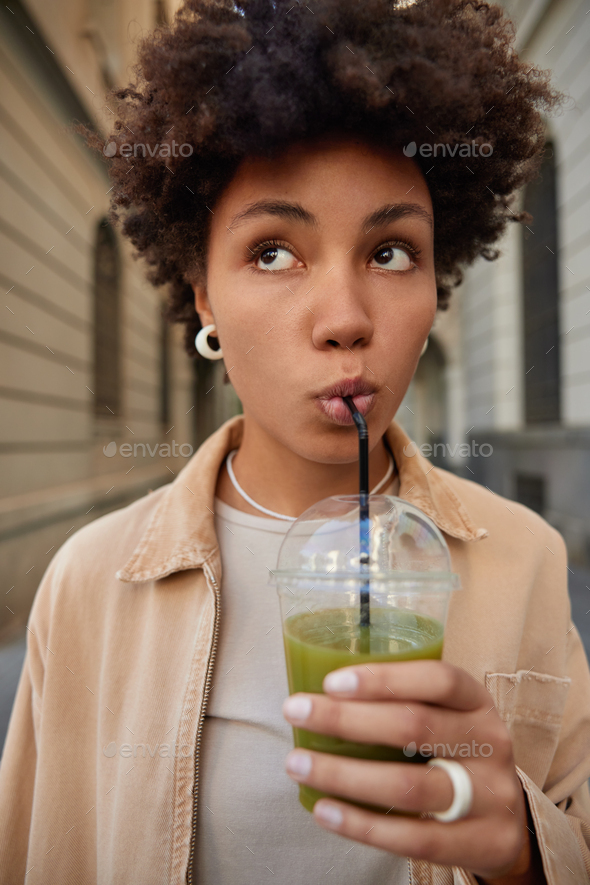 Vertical shot of thoughtful African American woman drinks natural green smoothie in plastic cup from