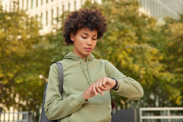 Horizontal shot of serious sporty woman checks time on smartwatch counts burned calories wears hoodi