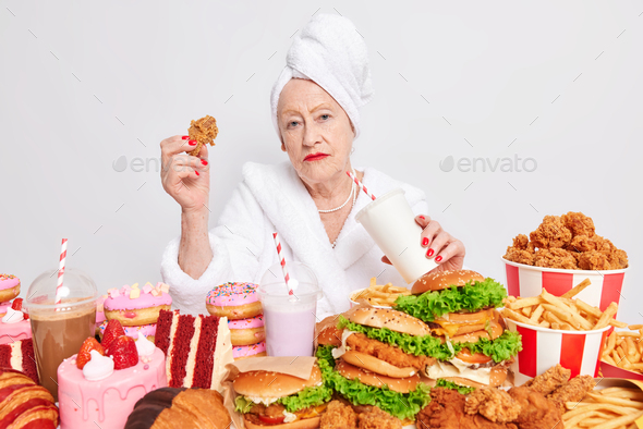 Fast food diet. Serious wrinkled senior woman drinks cocktail eats nuggets has overeating habits bre