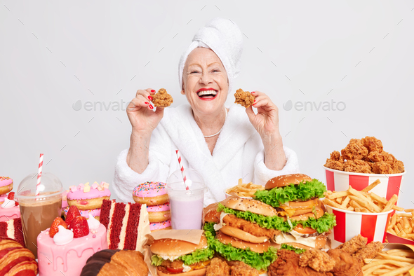 Cheerful wrinkled senior woman with red lips holds two nuggets eats cheat meal dressed in bathrobe s