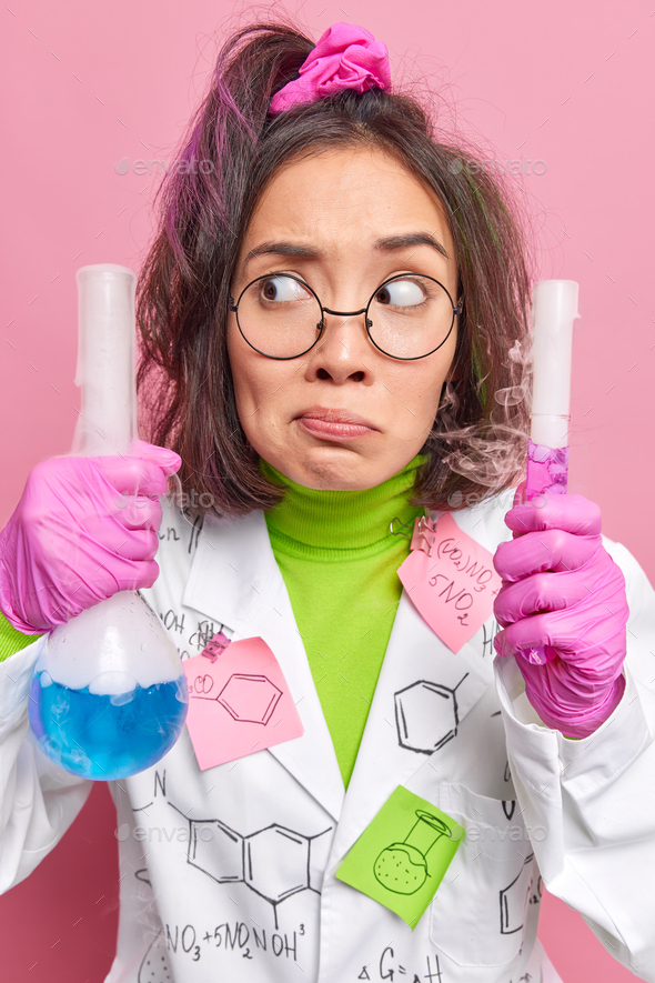 Confused puzzled female scientist holds two flasks with steaming liquid works in pharmaceutical labo